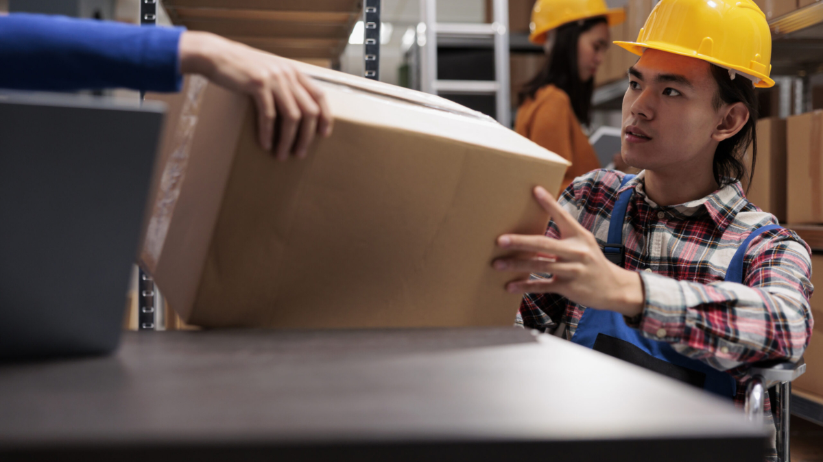 Storehouse order picker in wheelchair doing order fulfillment and taking cardboard box from coworker hands. Asian man with disability managing postal service operations in industrial warehouse